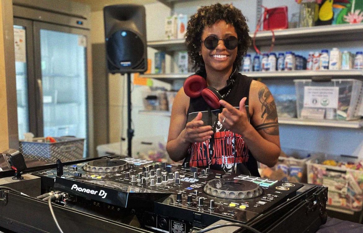 A person is smiling and standing behind a DJ setup with Pioneer DJ equipment in a room with shelves stocked with various cans and boxes. They are wearing sunglasses and holding headphones.