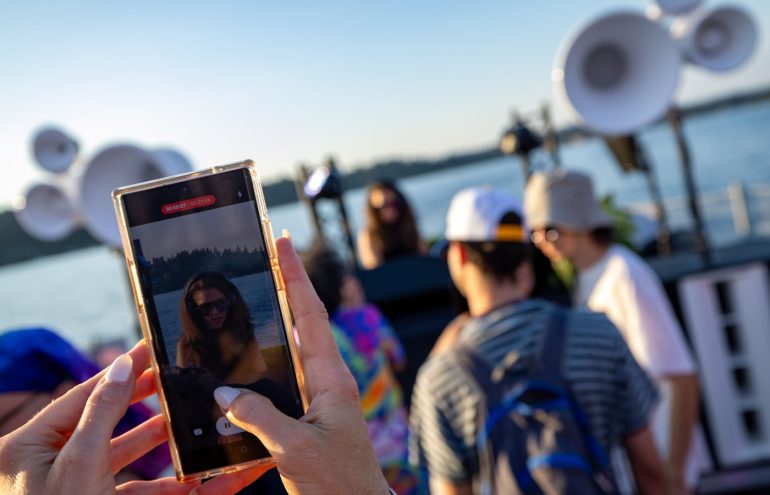 A person holds up a smartphone to take a photo of a DJ performing on a boat. The scene includes a sunny sky, water in the background, and several large speaker structures. Other individuals are nearby, facing the DJ.