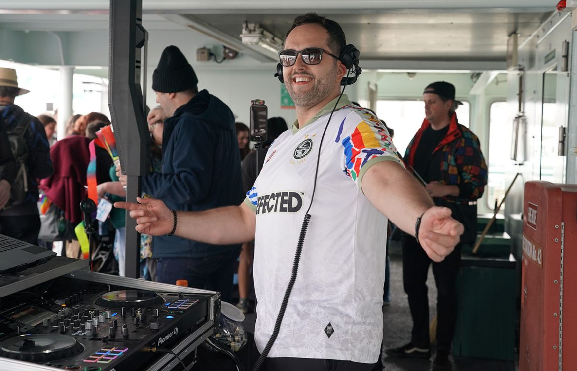 A person DJs on a ferry wearing headphones and a colorful sports jersey. They are smiling and standing beside DJ equipment. Other passengers are nearby, some wearing vibrant clothing.