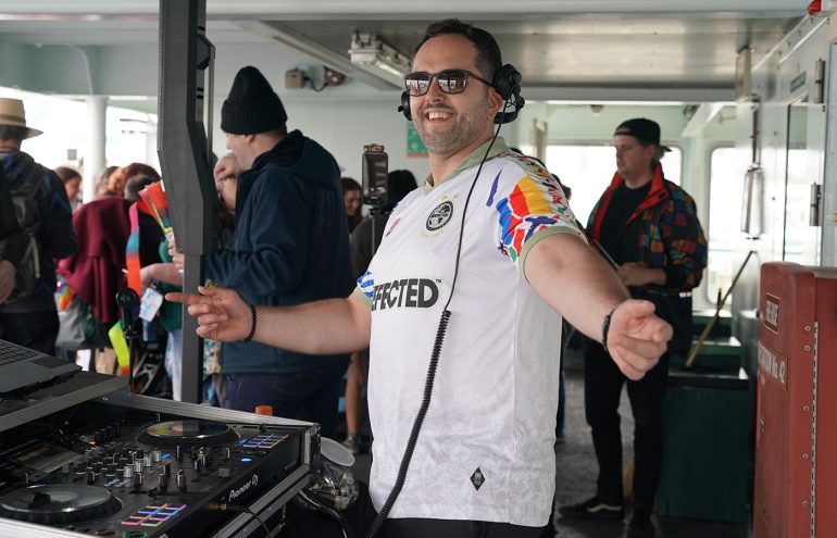 A person DJs on a ferry wearing headphones and a colorful sports jersey. They are smiling and standing beside DJ equipment. Other passengers are nearby, some wearing vibrant clothing.
